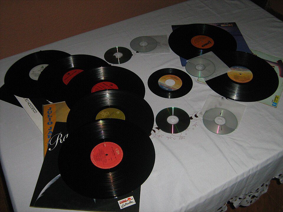 Several vinyl records and compact discs, scattered on a white surface, featuring various labels and colors, indicating a mix of old and new music formats, viewed indoors under dim lighting