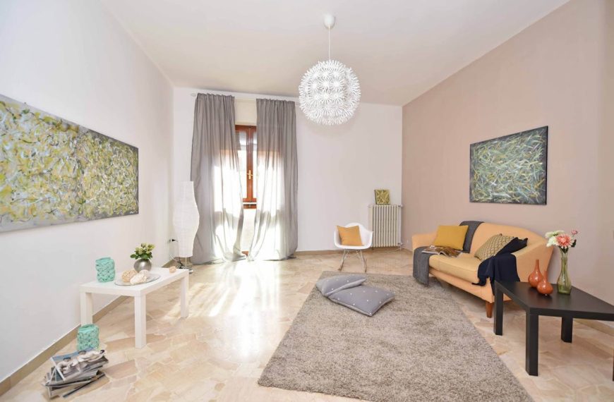 Modern living room interior, beige and white color scheme, soft gray rug, peach-colored sofa with cushions and throw blanket, black coffee table with vase and decor, white side table with candles and plant, abstract wall art, large window with sheer gray curtains