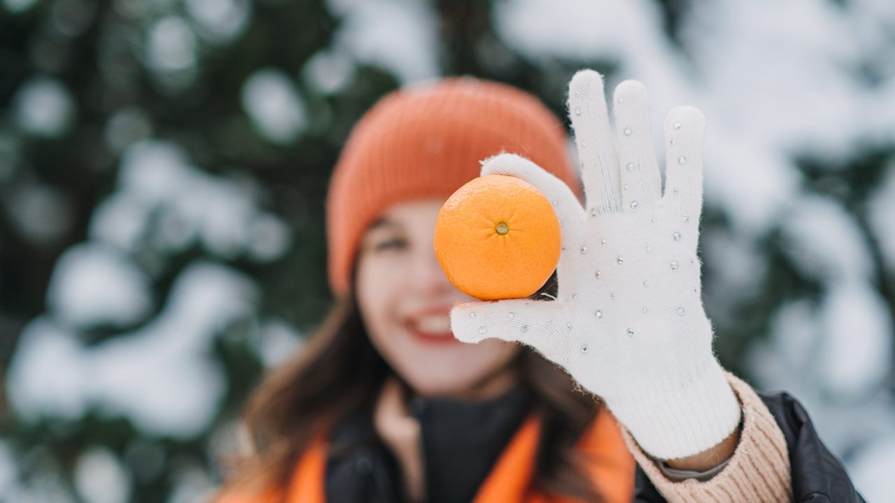 A woman outside in snow wearing winter gloves holding an orange up to the camera