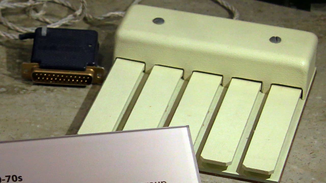 Close-up of the X-Y axis mouse prototype developed by Douglas Engelbart, featuring five rectangular buttons and a thick connector cable, displayed on a museum surface