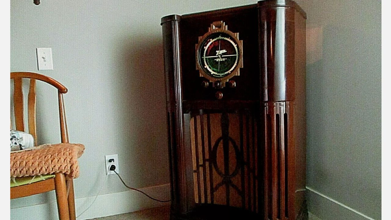 Vintage Zenith Stratosphere radio with polished wooden cabinet, large round dial, and ornate grille design, placed beside a wooden chair and plugged into a wall outlet