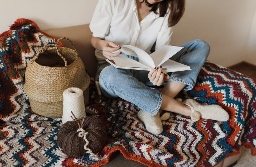 Woman in a white shirt and jeans, sitting cross-legged on a cozy couch, reading a book, surrounded by a colorful crochet blanket