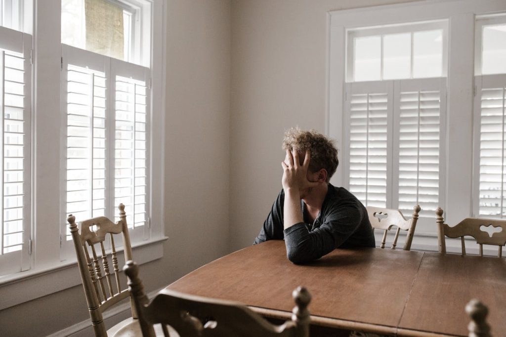 Man sitting alone at a wooden dining table in a bright room with white shutters, covering his face with one hand, appearing stressed or overwhelmed