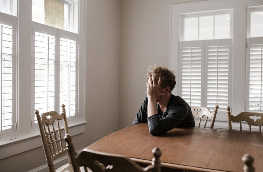 Man sitting alone at a wooden dining table in a bright room with white shutters, covering his face with one hand, appearing stressed or overwhelmed