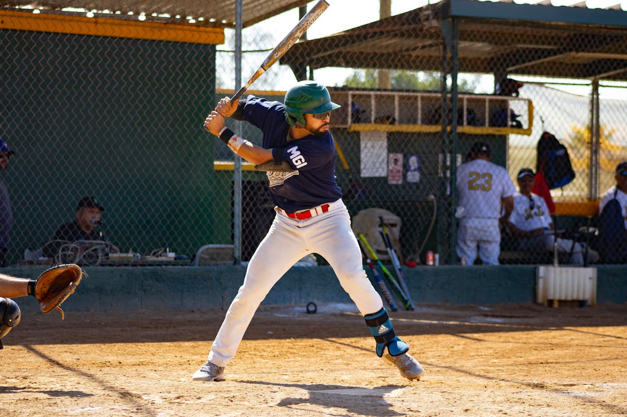 Baseball player in batting stance at home plate, wearing a helmet and gear, during a sunny game with teammates in dugout