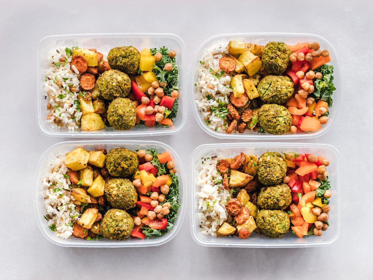Four meal prep containers filled with rice, roasted potatoes, carrots, chickpeas, kale, diced bell peppers, and green falafel
