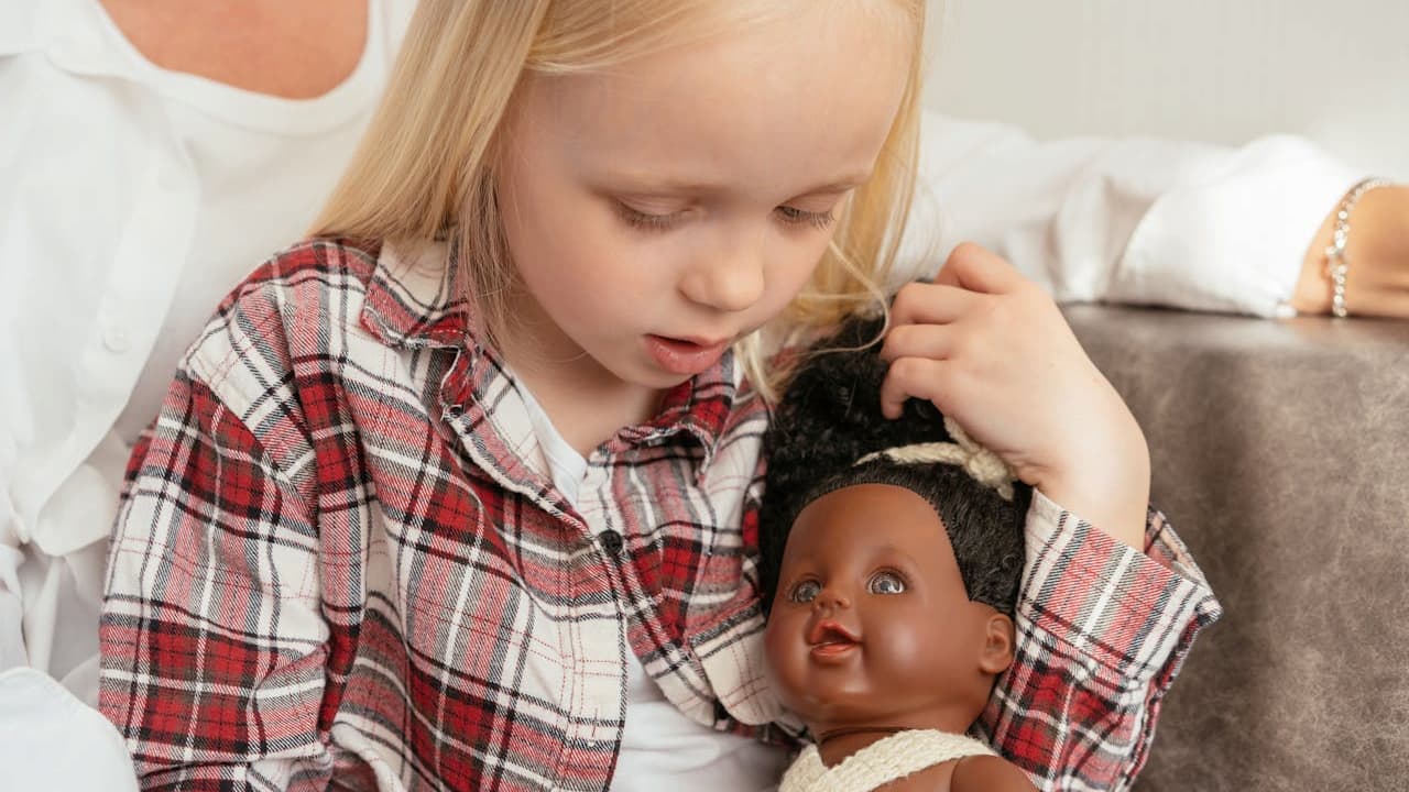 Young blonde girl in a plaid shirt holding a dark-skinned baby doll with curly black hair, sitting on a couch, woman in white shirt in background, soft lighting