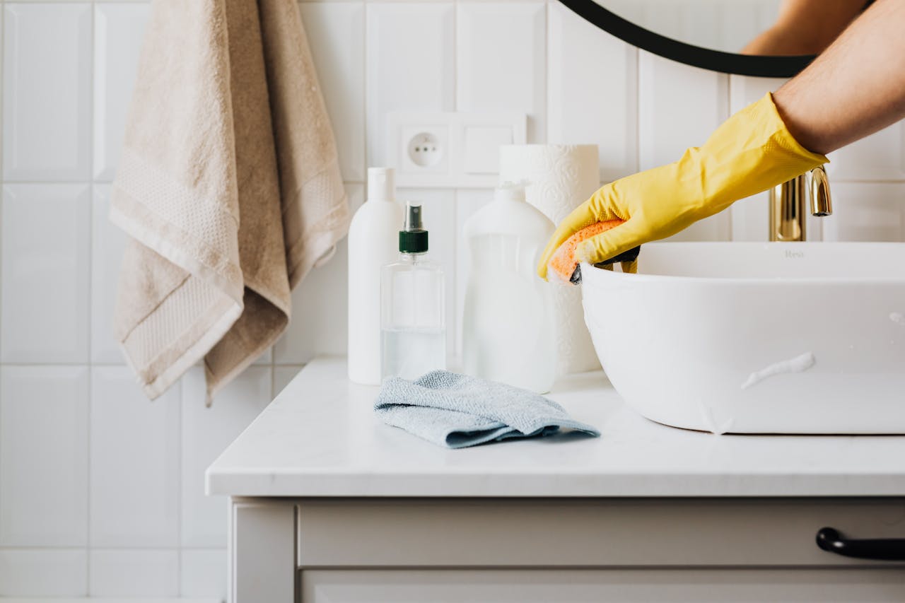 Person wearing a yellow rubber glove cleaning a white bathroom sink with an orange sponge, surrounded by various cleaning bottles