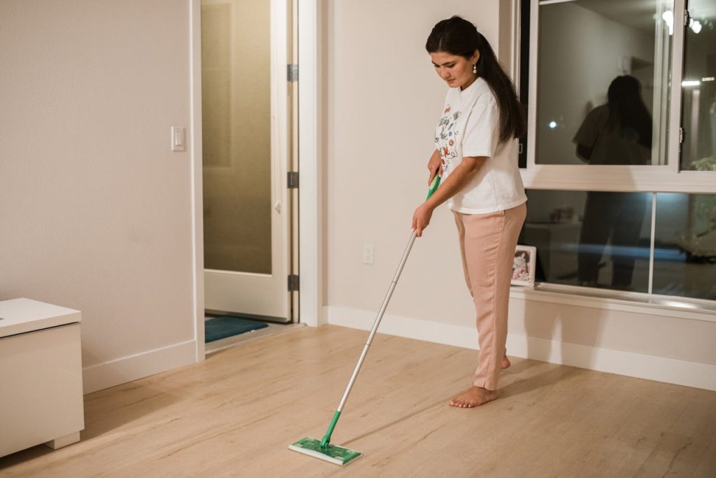 Woman mopping a wooden floor in a modern, bright living room with large windows and minimal decor