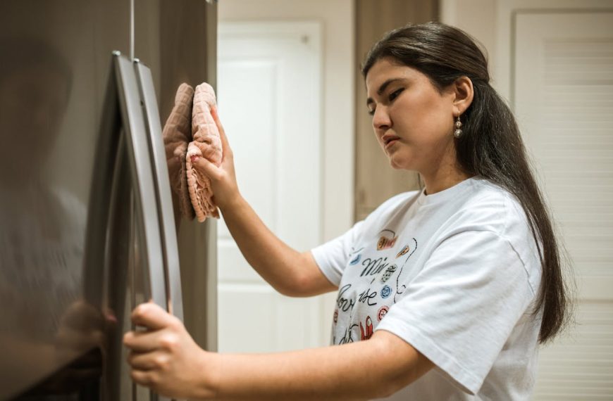 Woman in a white graphic t-shirt cleaning a stainless steel refrigerator with a pink cloth, focused expression, bright kitchen background