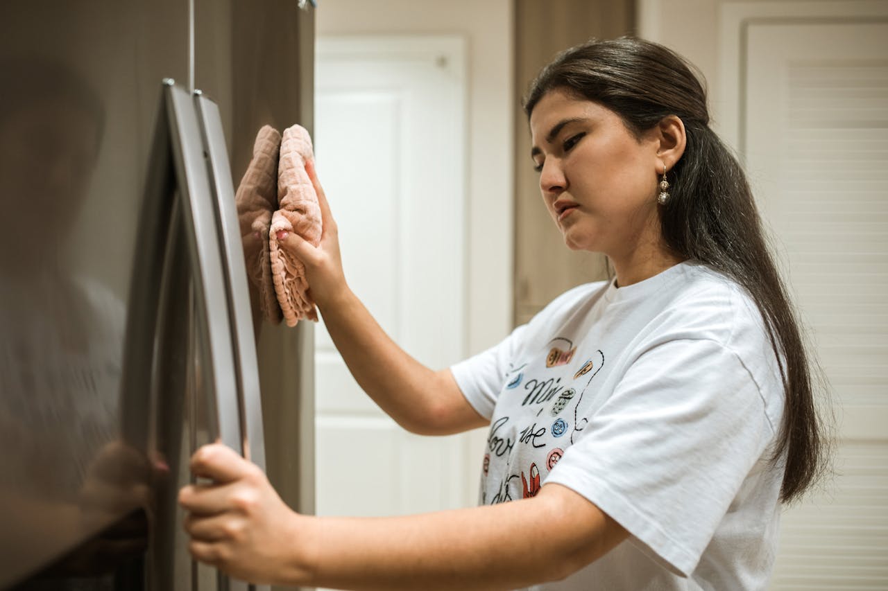 Woman in a white graphic t-shirt cleaning a stainless steel refrigerator with a pink cloth, focused expression, bright kitchen background