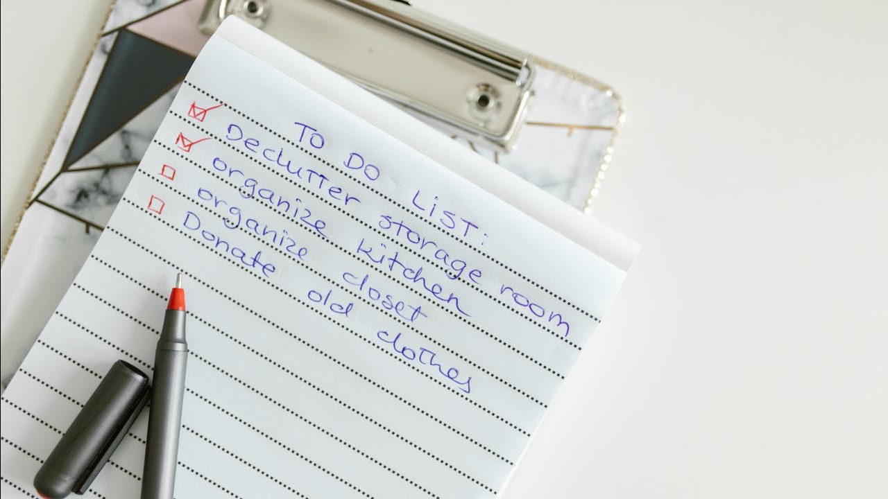 Notebook page with handwritten checklist in blue ink, black pen placed on the paper, smartphone and notebook partially visible underneath, white desk background