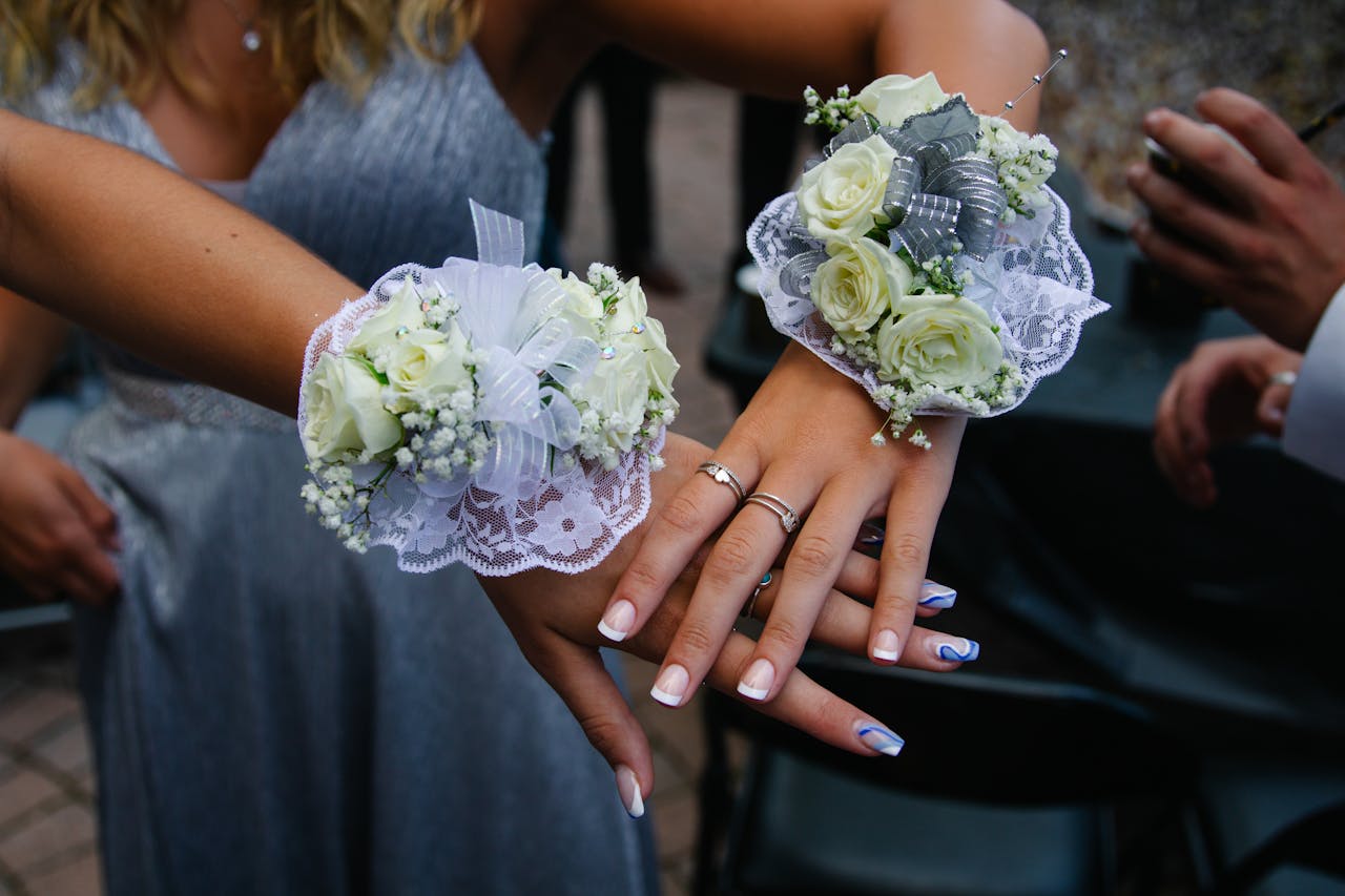 Two women wearing white rose corsages with lace and ribbon on their wrists, showing off manicured hands