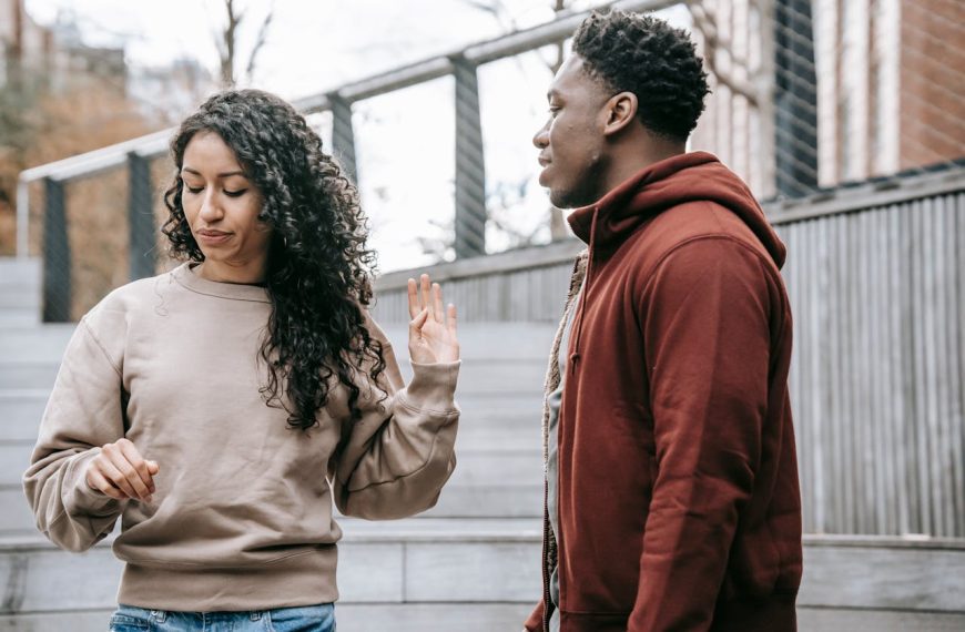 Woman raising her hand and turning away from a man during a conversation, both standing on outdoor steps, suggesting tension or disagreement