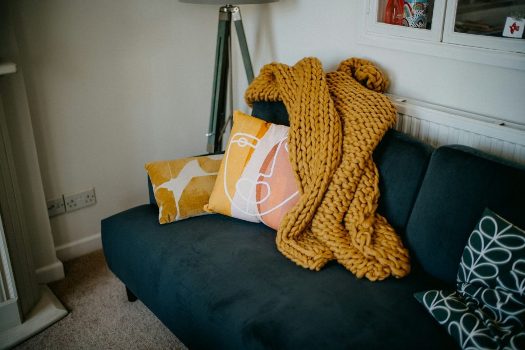 Dark blue couch, mustard yellow knitted blanket draped over armrest, two decorative pillows in yellow and white patterns, cozy and modern living room corner, white wall background, small radiator and lamp nearby, soft natural lighting