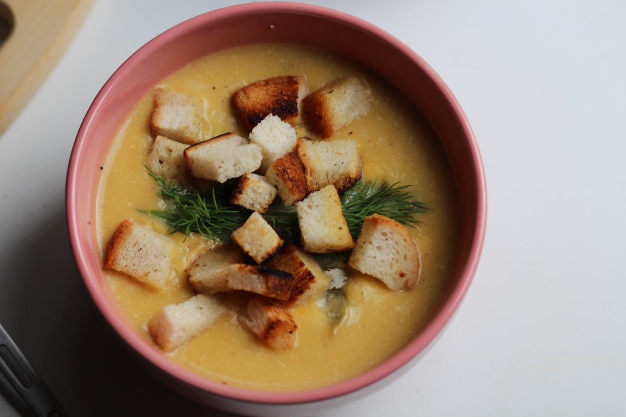 Creamy yellow soup in a pink bowl, topped with golden, crispy croutons and a fresh sprig of dill, on a white background