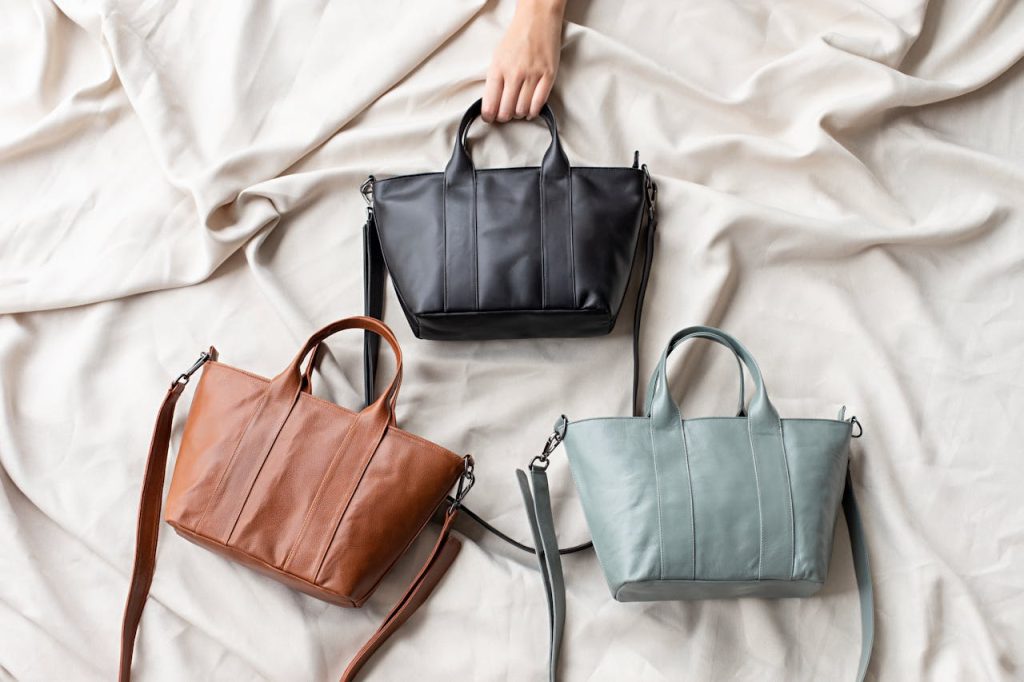Three leather handbags in black, brown, and light gray, arranged on a soft beige fabric with a hand reaching for the black bag