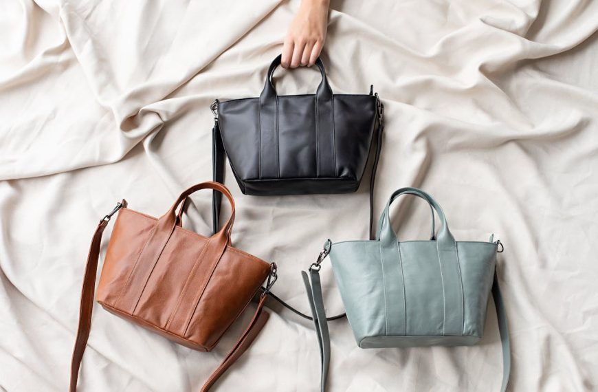 Three leather handbags in black, brown, and light gray, arranged on a soft beige fabric with a hand reaching for the black bag