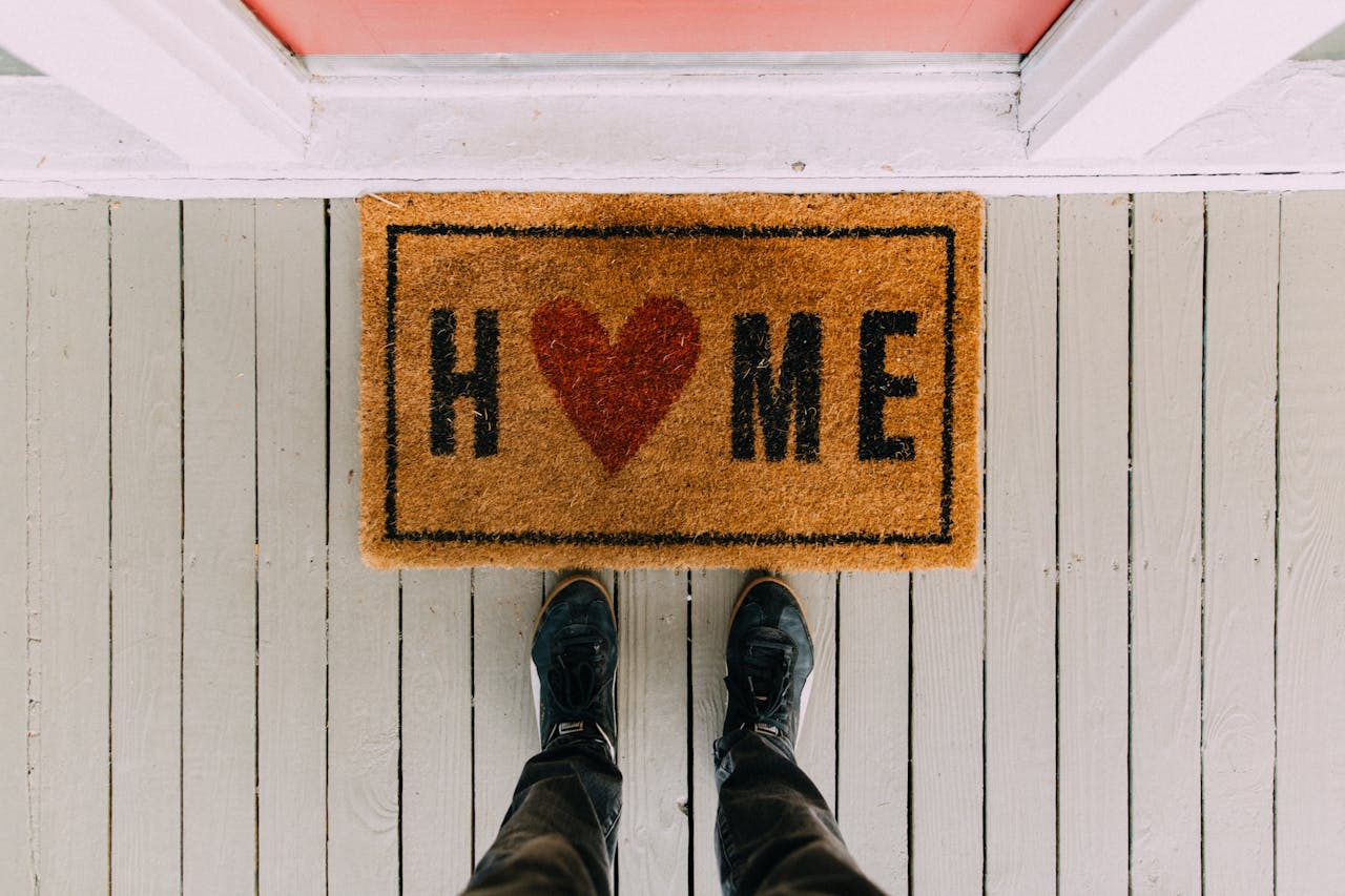 Brown doormat with the word "HOME" where the letter "O" is replaced by a red heart, placed on a light gray wooden porch