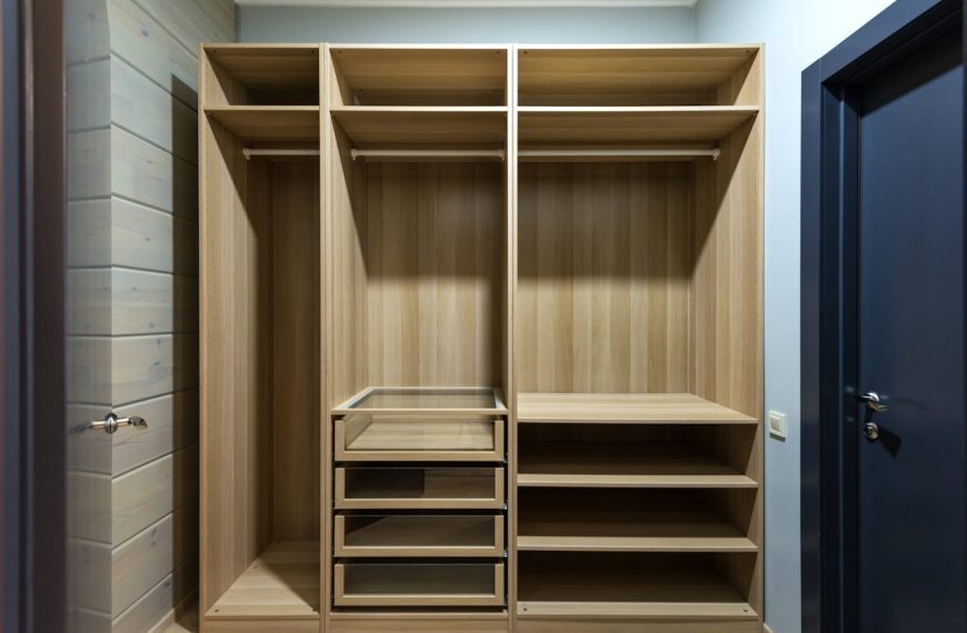 Empty wooden closet with open shelves, hanging rods, and glass-front drawers, surrounded by light wood paneling and dark door frames
