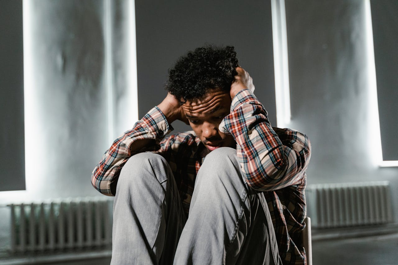 Man sitting curled up on a chair with hands over his ears, looking distressed in a dimly lit room with white walls and vertical windows