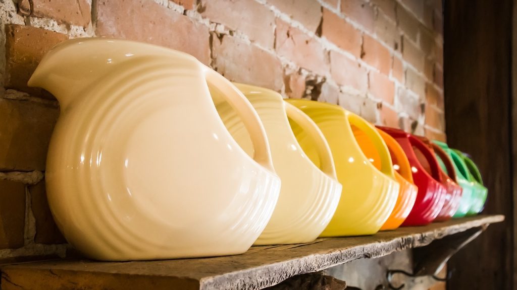 An angled view of a colorful row of fiesta-ware water pitchers, aligned with handles out, on a rustic wooden shelf with brick wall background