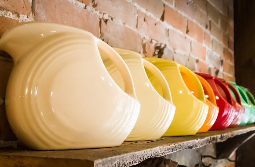 An angled view of a colorful row of fiesta-ware water pitchers, aligned with handles out, on a rustic wooden shelf with brick wall background