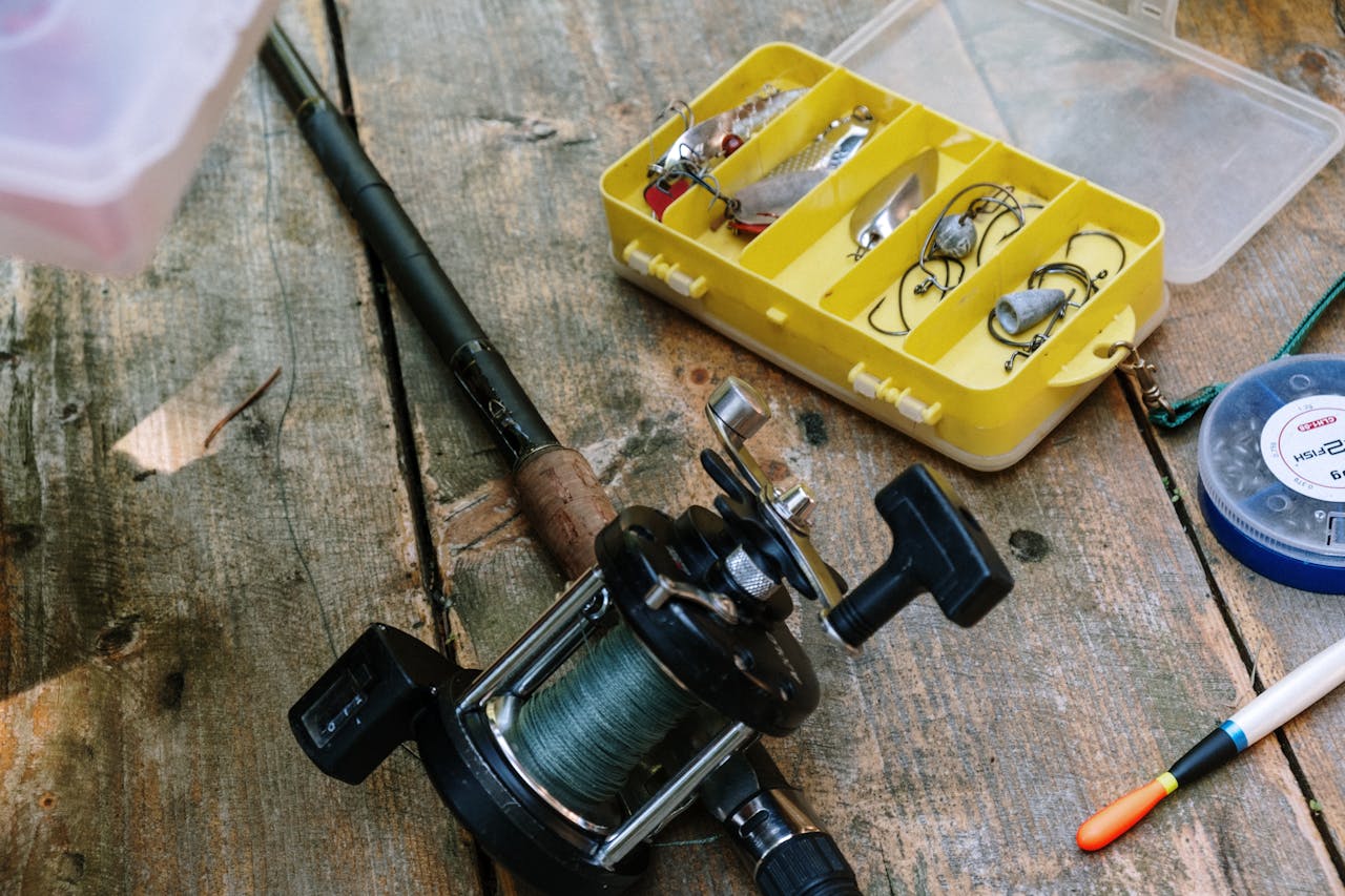 Fishing gear on a rustic wooden surface, including a fishing rod with a black reel, a yellow tackle box with hooks