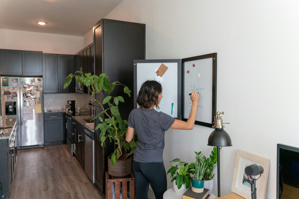 Person writing on a wall-mounted whiteboard, modern apartment kitchen in the background, indoor plants nearby, wooden floor, organized and minimalist living space.