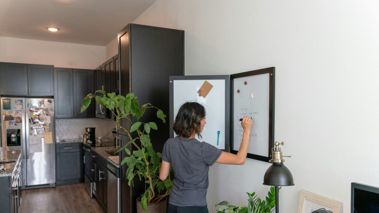 Person writing on a wall-mounted whiteboard, modern apartment kitchen in the background, indoor plants nearby, wooden floor, organized and minimalist living space.