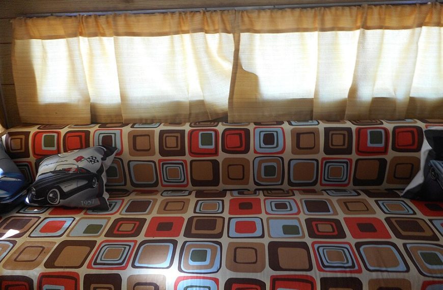 Long patterned sofa with multicolored square designs, flanked by armrest pillows, positioned beneath a window with cream curtains, sunlight filtering through the fabric