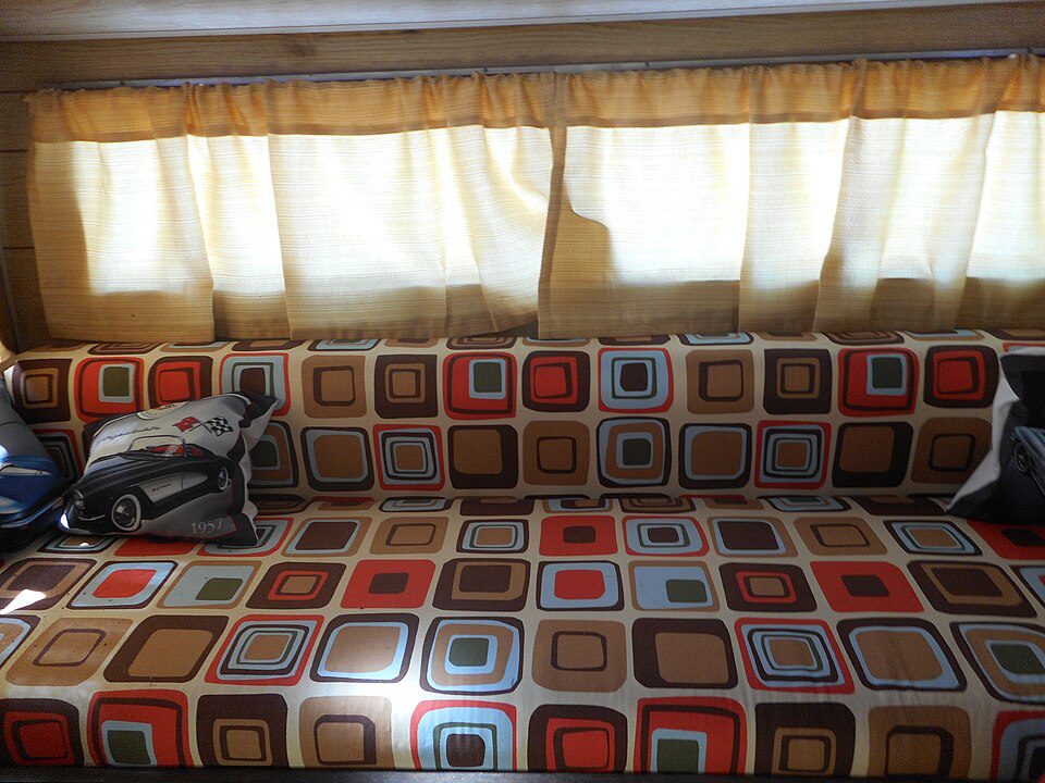 Long patterned sofa with multicolored square designs, flanked by armrest pillows, positioned beneath a window with cream curtains, sunlight filtering through the fabric