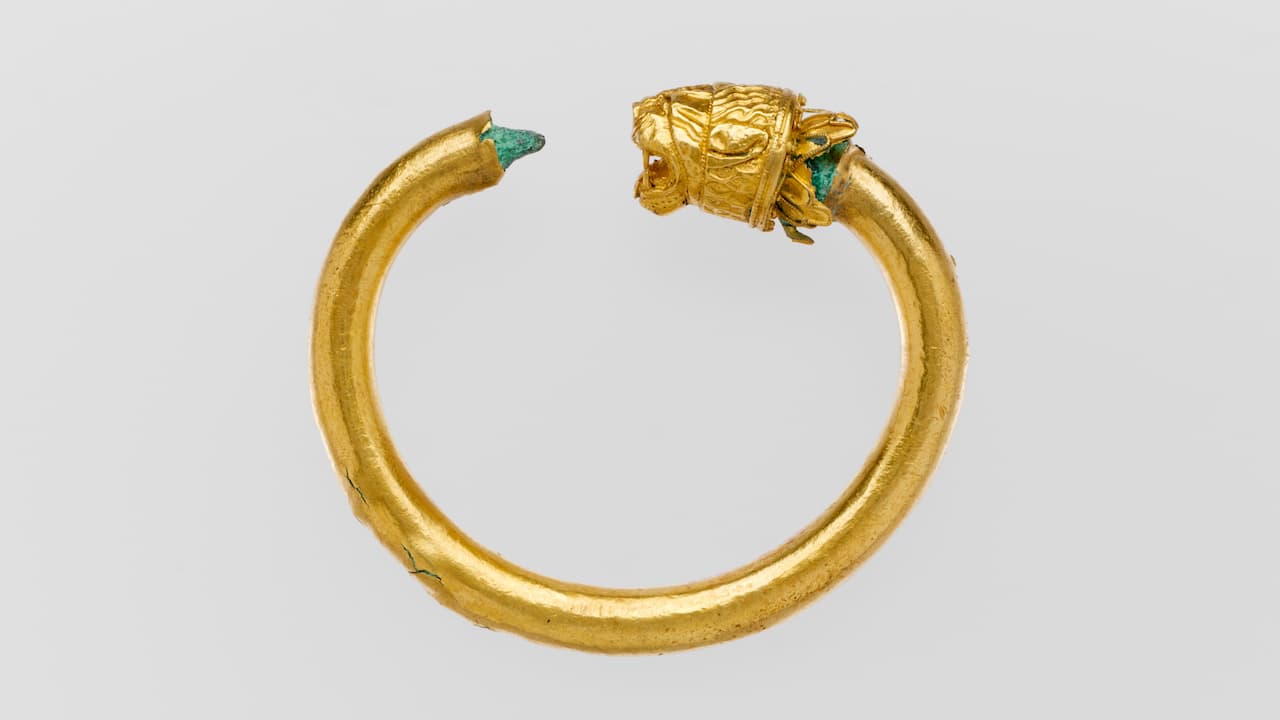 Gold bracelet with a lion-head design, open-ended, green corrosion visible at the tip and near the decorative head, smooth circular band, set against a plain light background.