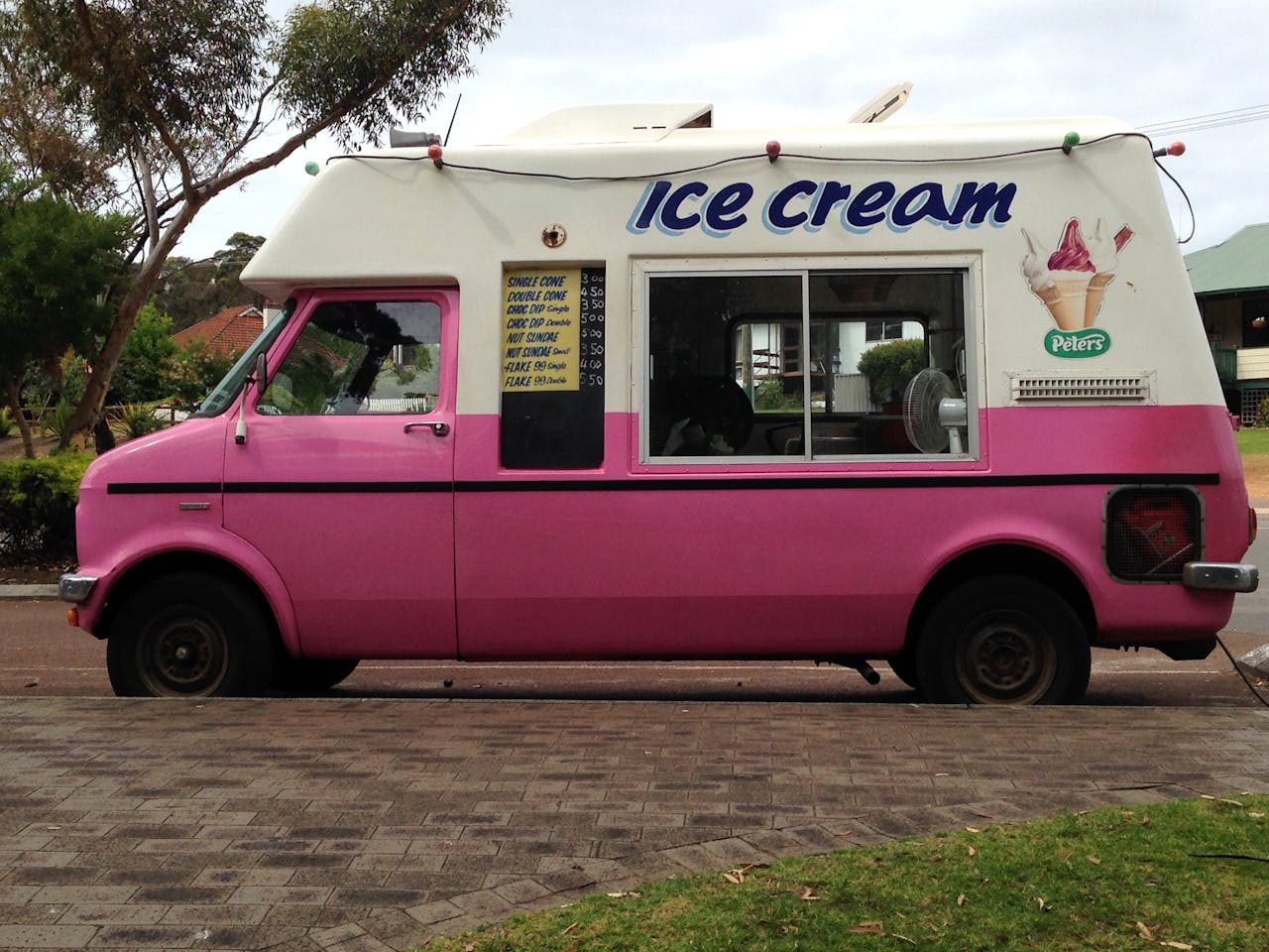 Classic pink and white ice cream truck parked on a suburban street, decorated with colorful string lights and a menu board displaying ice cream cone options