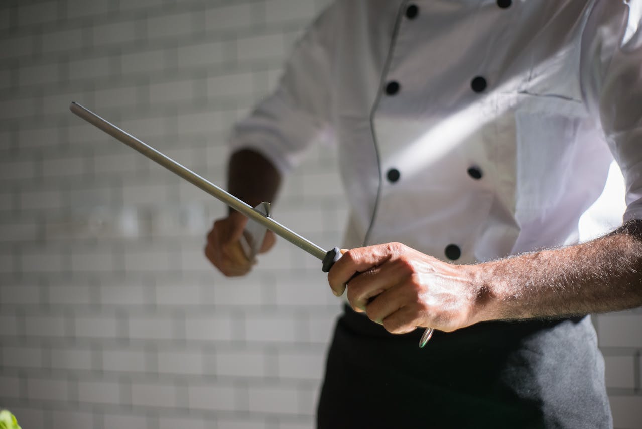 Chef sharpening a knife with honing steel, dressed in white uniform with black buttons, kitchen background, tiled wall