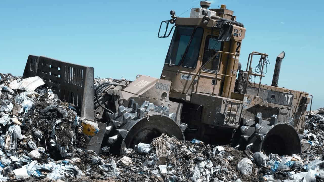 A heavy-duty landfill compactor vehicle crushes piles of mixed waste and garbage at a landfill site under a clear blue sky