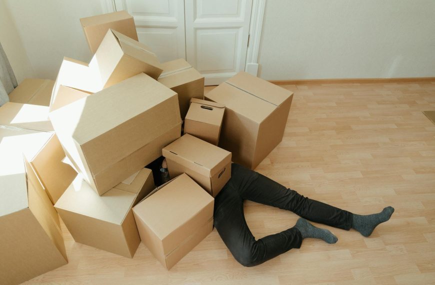 person lying on the floor, covered by cardboard boxes, light wood flooring, white wall and door in background, scene suggests moving or unpacking, legs visible wearing black pants and shoes