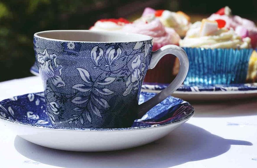 Blue and white floral-patterned teacup and saucer, part of a fine china set, placed on a table beside decorated cupcakes in colorful wrappers