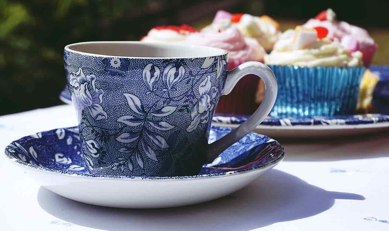 Blue and white floral-patterned teacup and saucer, part of a fine china set, placed on a table beside decorated cupcakes in colorful wrappers