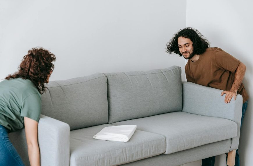 A light gray modern sofa with a white book or tablet resting on the seat cushion. The edge of a person with dark curly hair is partially visible behind the sofa's right side.