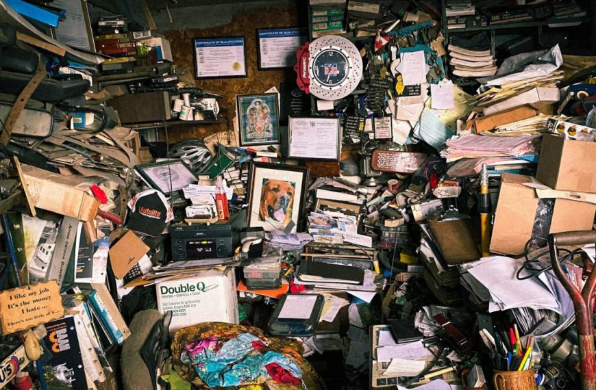 Extremely cluttered room filled with stacked papers, cardboard boxes, tools, books, framed photos, certificates, and miscellaneous objects covering every surface, walls, and shelves, with a red and white wall clock and a dog portrait at the center