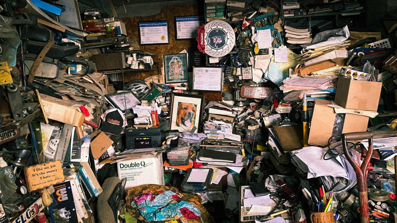 Extremely cluttered room filled with stacked papers, cardboard boxes, tools, books, framed photos, certificates, and miscellaneous objects covering every surface, walls, and shelves, with a red and white wall clock and a dog portrait at the center