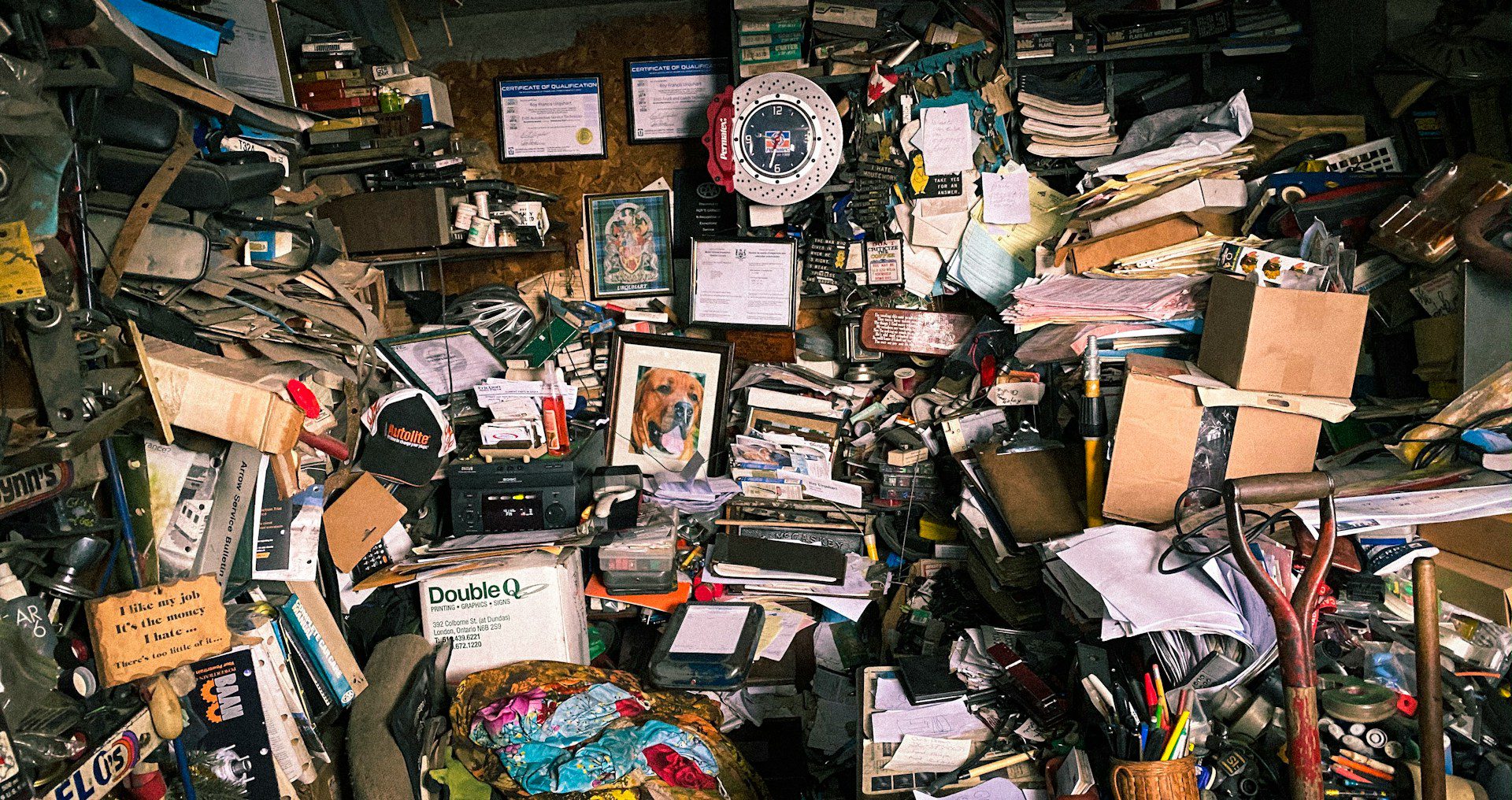 Extremely cluttered room filled with stacked papers, cardboard boxes, tools, books, framed photos, certificates, and miscellaneous objects covering every surface, walls, and shelves, with a red and white wall clock and a dog portrait at the center