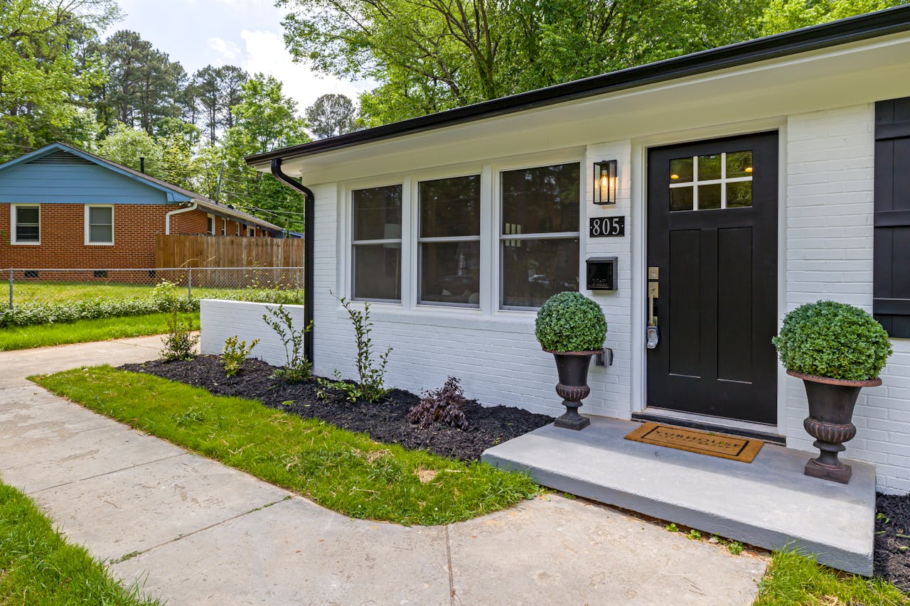 Front of a modern white brick house with black door, house number 805, two potted shrubs, and a new concrete walkway