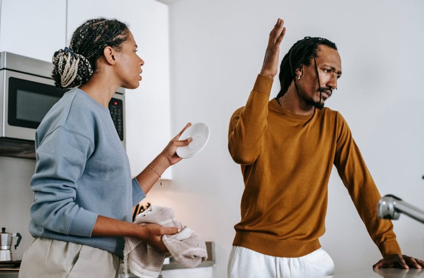 Woman holding a plate and towel while arguing with a man who raises his hand in frustration in the kitchen