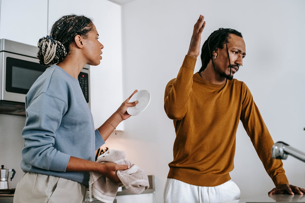 Woman holding a plate and towel while arguing with a man who raises his hand in frustration in the kitchen