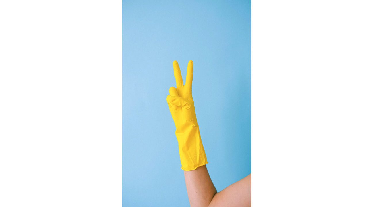 Arm wearing a yellow cleaning glove making a peace sign against a light blue background