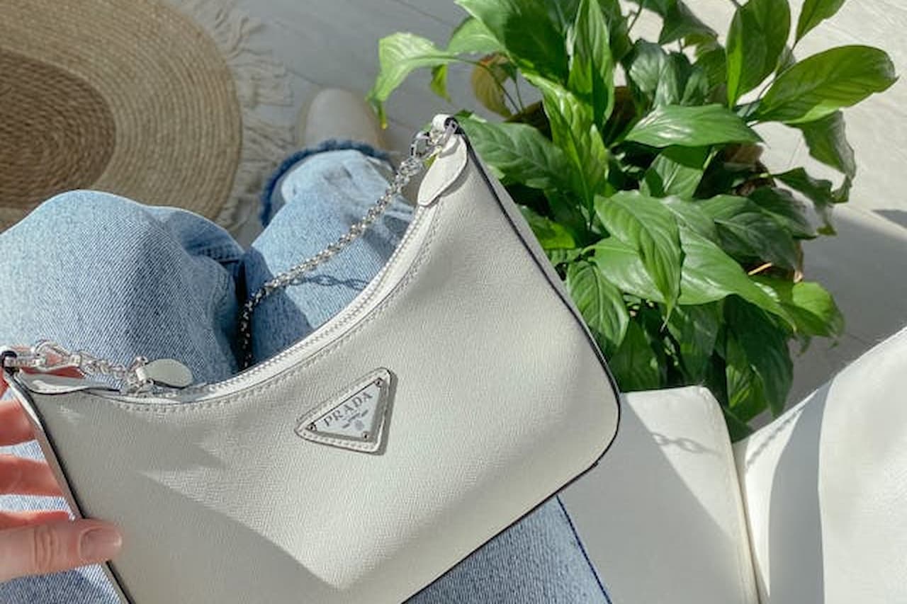 White designer handbag, triangular metal logo, resting on denim jeans, green potted plant in background, bright natural lighting