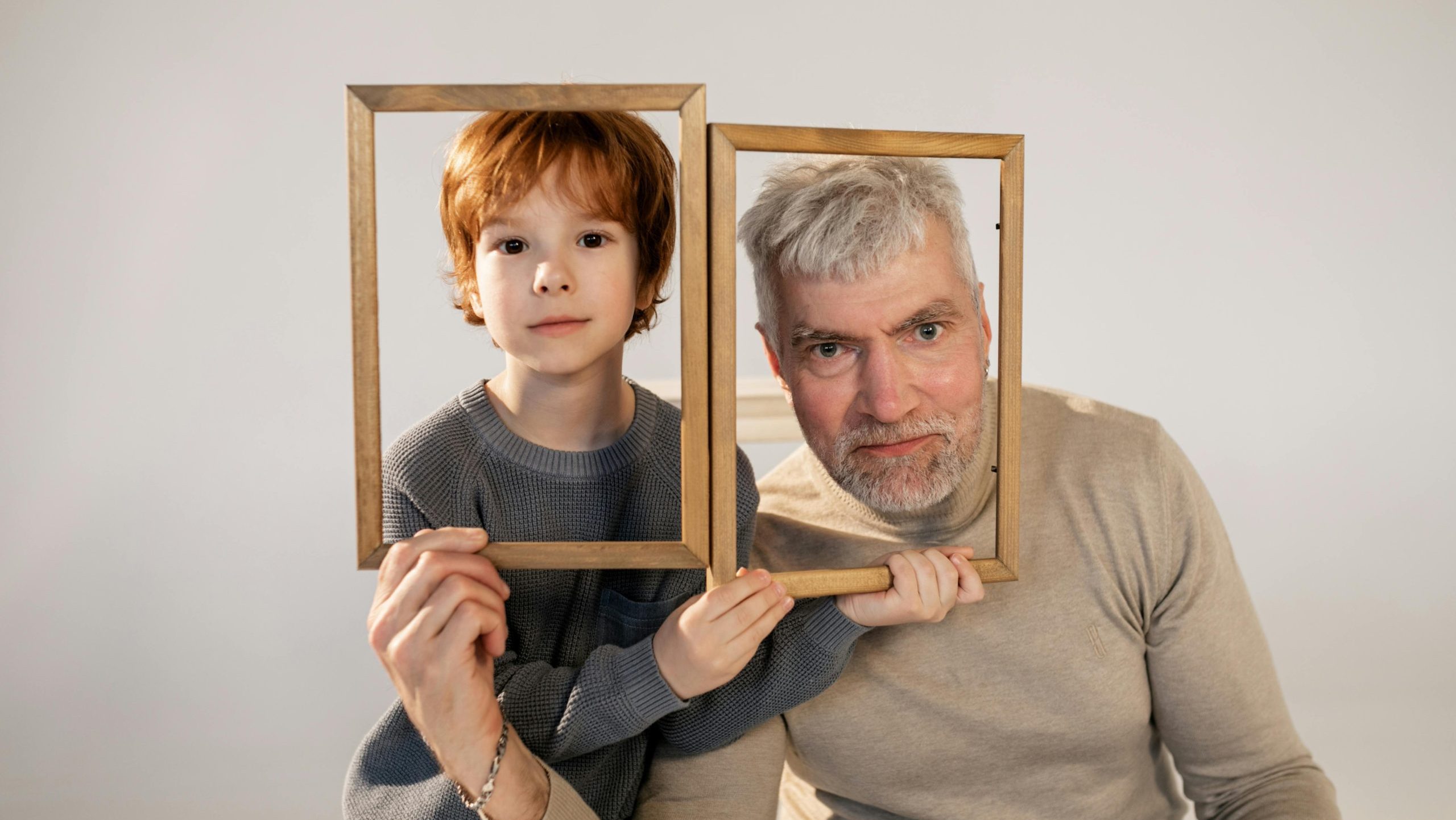Young boy and older man sitting closely together, each holding a wooden photo frame in front of their faces, both looking directly at the camera, neutral background, playful and warm family portrait concept