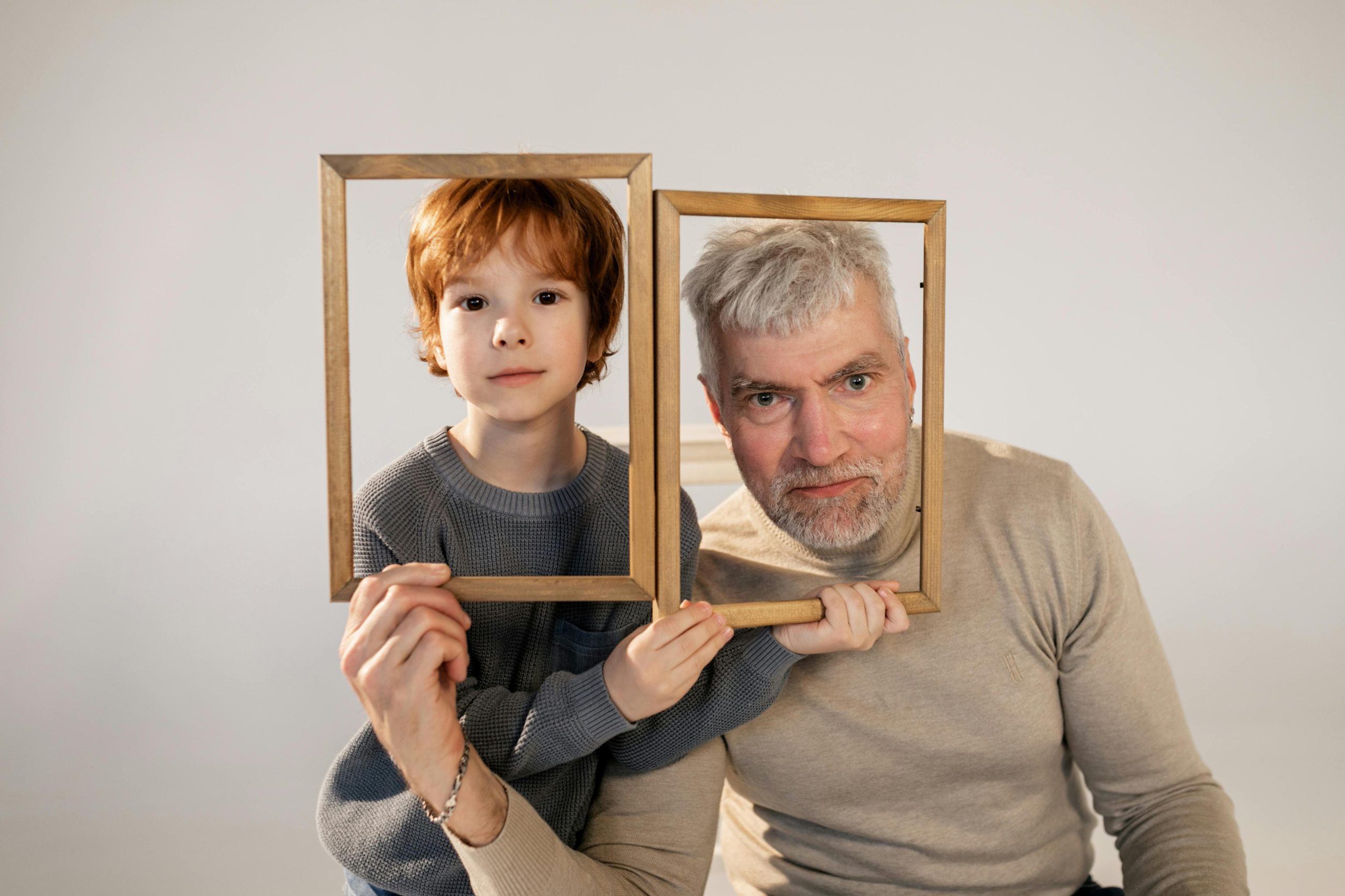 Young boy and older man sitting closely together, each holding a wooden photo frame in front of their faces, both looking directly at the camera, neutral background, playful and warm family portrait concept